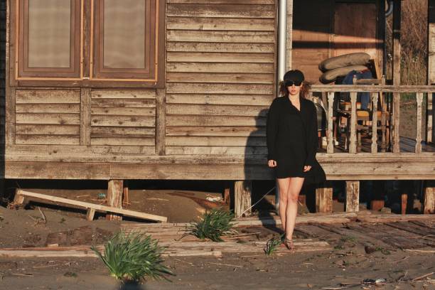 Beautiful blond haired girl wearing black flat cap, dress and sunglesses standing in front of her wooden beach house porch