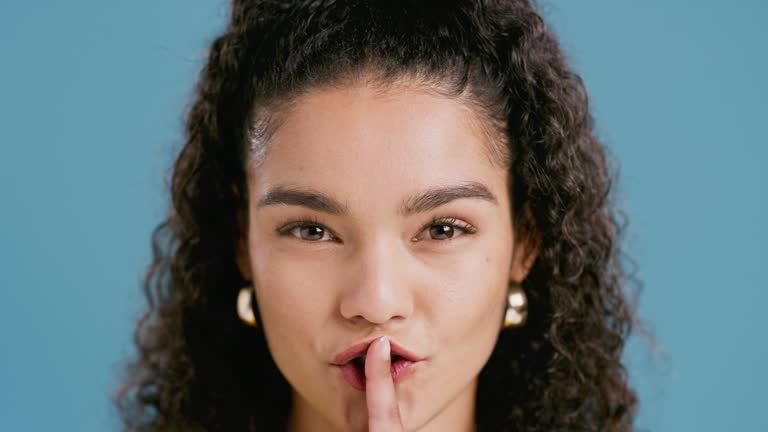 Closeup, woman and studio with mouth in secret for noise, privacy and confidential information with finger sign. Person, emoji and quiet for mystery or portrait with hand gesture on blue background