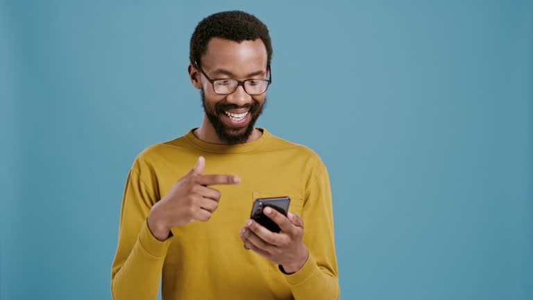 Face, cellphone and happy man with pointing in studio for bonus on blue background for notification or prize. Smile, portrait and african model for winner and celebration in online competition mockup