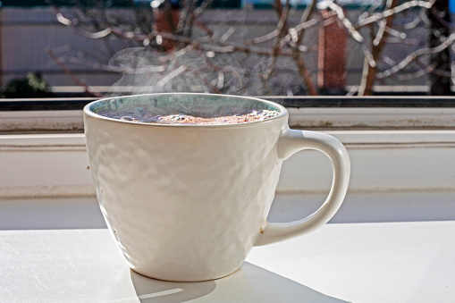 white cup of hot drink with tea or coffee stands on the sill near the window. Day off