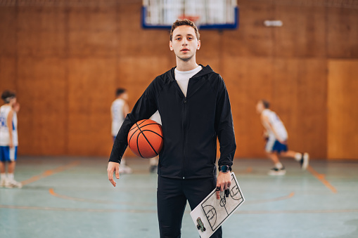 Portrait of a basketball coach standing on court with a ball and clipboard in his hands and looking at the camera. In a blurry background is his junior basketball team practicing basketball on court.