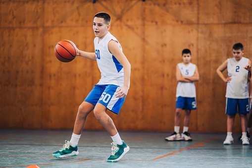 Portrait of a junior basketball player in action dribbling a ball on basketball court during the training. In a blurry background are his teammates watching him. An active basketball kid in motion.