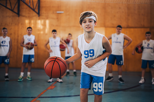 Dynamic portrait of a junior basketball player dribbling a ball and practicing basketball on training at indoor court. In a blurry background is his team watching. A boy in action with basketball.