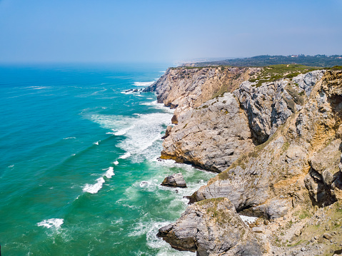 Rocky cliffs adorned with green vegetation overlook a secluded sandy beach where turquoise sea waves crash, captured from an aerial perspective.