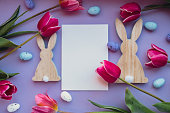 White empty envelope copy space Easter eggs and wooden bunnies with pink tulips on violet pastel background. Greeting card Postcard. Flat lay composition top view. Decoration minimalist modern design template. Floral layout Concept of creative spring