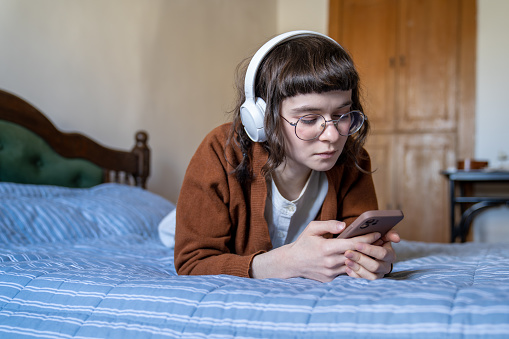 Relaxed music lover teen girl listening to music in headphones lying in bed at home. Pensive teenage schoolgirl using smartphone in earphones. Music fan enjoying leisure time pastime with mobile phone