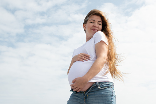 Happy Caucasian pregnant woman holding her belly on the sky background. white t-shirt and jeans. Third trimester. Happy pregnancy concept. Maternity care
