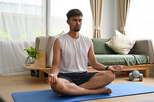 Healthy Asian man practicing yoga at home, sitting in lotus pose to meditation. Healthy lifestyle, meditation concept.