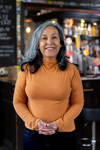 Three-quarter-length portrait of a mature woman smiling, looking into the camera with her hands clasped in front of her. She is wearing smart casual attire. The bar/restaurant is located in Hexham.