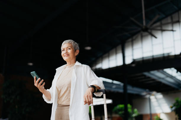 Asian senior woman business travel An Asian Chinese businesswoman with luggage using smartphone to order a ride during her trip business journey stock pictures, royalty-free photos & images