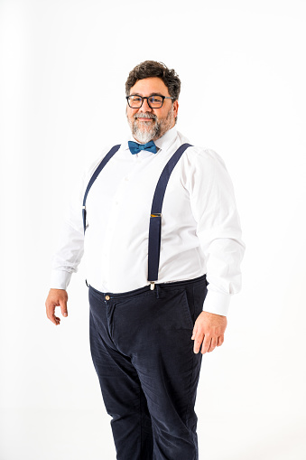 A cheerful, bearded middle-aged man stands confidently against a white backdrop looking at camera, dressed in a crisp white shirt with blue suspenders and a matching bow tie and he wears glasses
