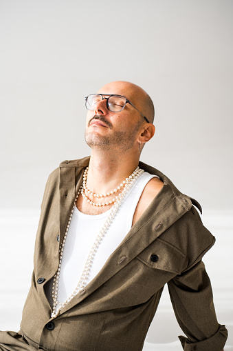 A queer person is sitting on the ground while wearing a pearl necklace in a studio fashion portrait.