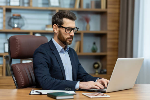 istock Focused male in eyeglasses doing computer work while sitting in leather chair by wooden desk in workplace. Attentive careerist checking terms of conditions to applying for heading new projects 2027814578