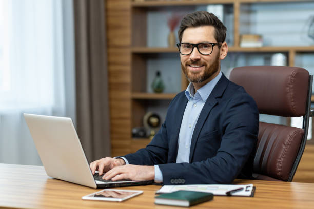 istock Smiling attractive man in stylish suit holding hands on pc keyboard and confidently looking at camera. Successful employee feeling satisfied about working in company and personal career achievements 2027815087