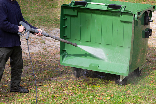 A municipal service worker treats a garbage container with a disinfectant solution. Washing the garbage container.