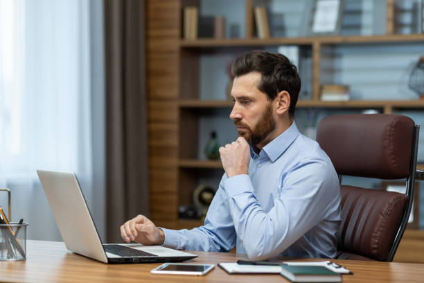 istock Focused bearded man looking attentively at portable computer screen and holding hand under chin in workspace. Small business owner checking monthly statements online and accounting bills 2027821963