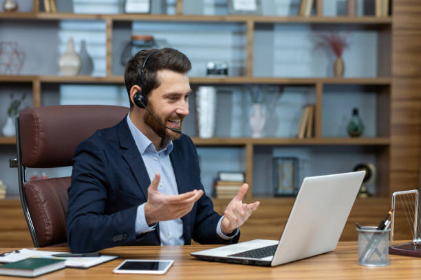istock Positive man in wireless headset with micro hosting video conference while sitting by desktop with computer. Millennial businessperson using modern technology for online communication with partners 2027822172