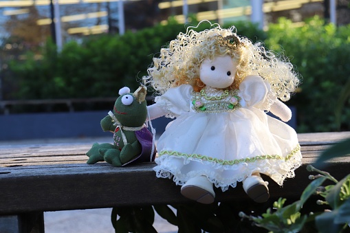 A doll sitting on a bench with bushes in the background.