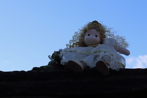 The image shows a stuffed doll placed on a ledge against a backdrop of the sky with clouds in the background.