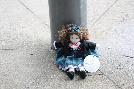 The image is of a doll that is placed outdoors, sitting in front of a column that has lines going outwards.
