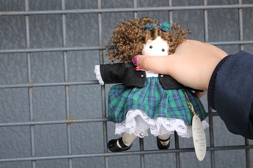 The image is of a person holding a stuffed doll outdoors. The person's clothing, the stuffed toy, a wall, and the ground are visible in the outdoor setting.