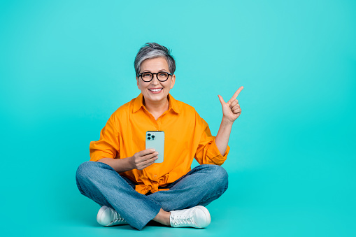Full body photo of senior smiling woman business project manager indicate finger mockup with phone isolated on aquamarine color background.