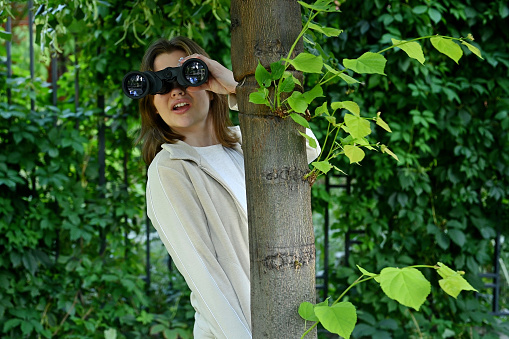 Close-up of a young woman watching someone with binoculars while hiding behind a tree
