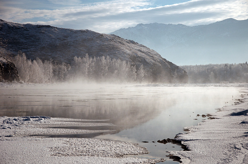 A serene winter river enveloped in fog with majestic mountains, trees in frost, and a cloudy sky in the background, creating a picturesque natural landscape