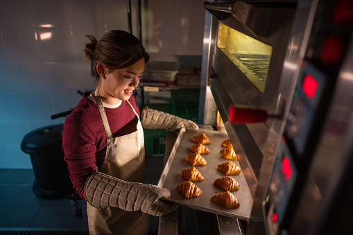 An Asian baking trainee is holding a baking sheet filled with buns in a commercial kitchen at a cooking school.