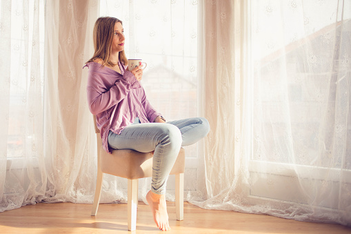 The woman is sitting on a chair and holding a mug . The woman is drinking tea and enjoying the view from her window.