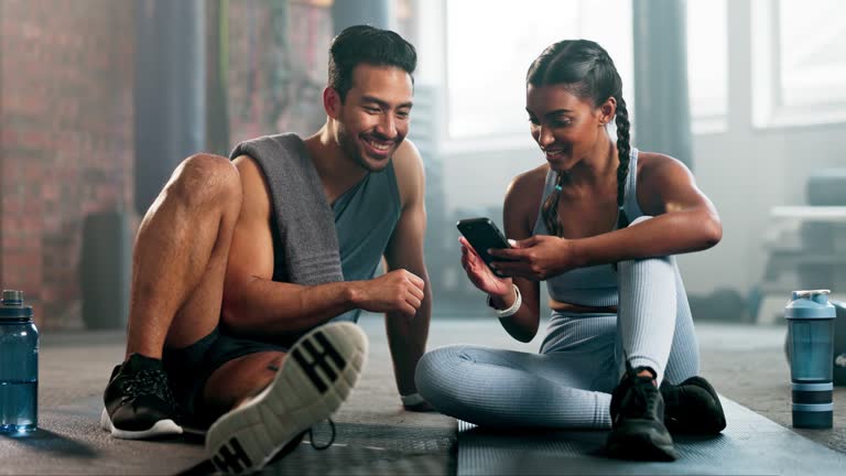 People, phone and talking fitness at gym for training or exercise app on internet. Asian man and indian woman or workout friends with smartphone for good review or ok sign for personal trainer