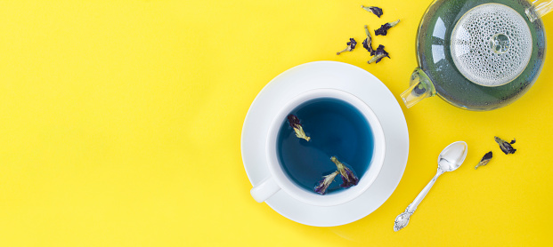 Blue flower tea in the white cup and glass teapot on the yellow background. Top view. Copy space.