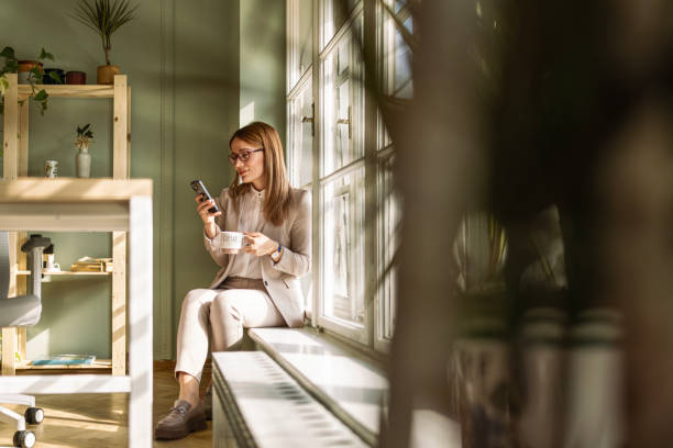 istock Businesswoman Relaxing on Window Ledge with Smart phone Text Messaging 2028150871