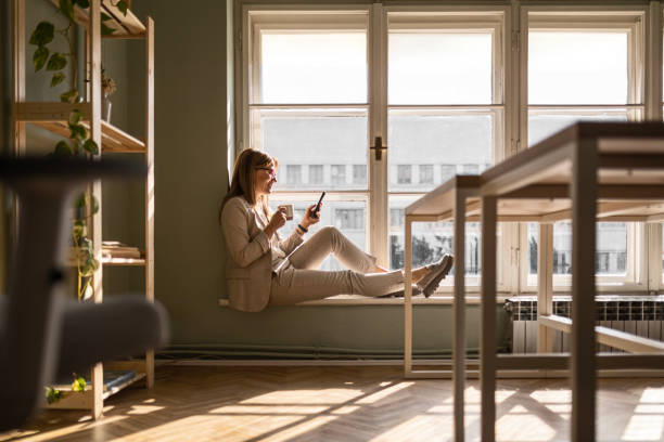 istock Businesswoman Relaxing on Window Ledge with Smart Phone and coffee cup 2028150876