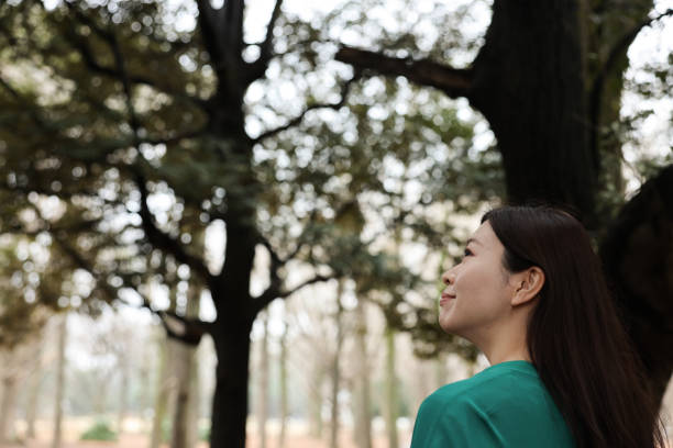 Beautiful smiling happy woman touching a green branches with leaves of a young maple tree in the forest in sunny summer weather. Girl rest, breathing air deep in fashion sport clothes in the woods