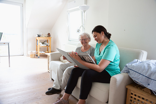 Senior woman sitting on sofa looking at photo album with female nurse, sharing memories,photos are black and white, a white cat with black spots is sitting besides them