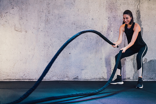 Shot of a strong beautiful young woman working out with battle ropes in a cross training gym. Sporty woman doing cross exercising with ropes in workout gym. Determined girl using battle rope while doing physical training.