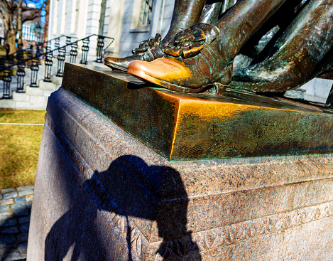 Cambridge, Massachusetts, USA - February 19, 2024: Shadow of a photographer holding a camera up to photograph the shoe toe of the John Harvard statue, a popular photographic  subject for tourists. People often rub the toe of John Harvard's left shoe for luck, hence its shiny gold color. The John Harvard (1607-1638) statue inscription is \