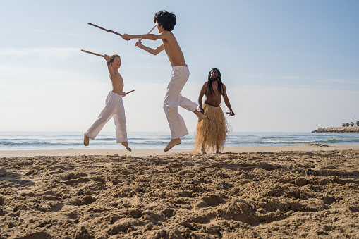 Boys in white pants practicing capoeira (Brazilian martial art that combines elements of dance, acrobatics and music) on the beach in front of their teacher's gaze