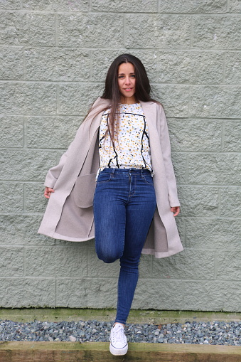 A Mexican woman leaning against a brick wall with one leg up. She is wearing long, brown, straight hair, an open coat, pattern shirt, jeans and white shoes.