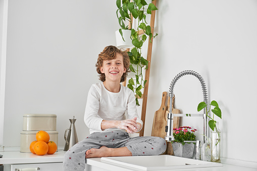 Barefoot boy in sleepwear sitting on kitchen counter and washing hands with soap while looking at camera with happy smile