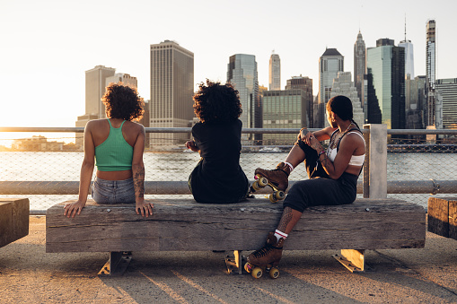 Three women with roller skates taking a break in front of New York City skyline at sunset.
