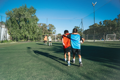 rear view on two boys from opponent teams walking embraced after match on pitch