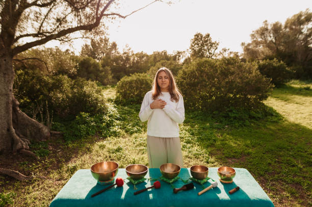 mulher adolescente bonita com olhos fechados de pé com as mãos em seu coração antes de fazer meditação usando várias tigelas de canto tibetano em seu jardim - spirituality hand on heart meditating women - fotografias e filmes do acervo