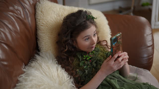 A girl in a beautiful green dress looks at the screen of a mobile phone, smiles, laughs and grimaces, lying on a sofa in a modern living room. A girl watches entertainment content from the Internet.