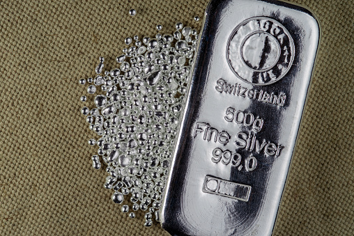 A silver bar weighing 500 grams  and a pile of silver grains on the background to the coarse texture of the textile.
