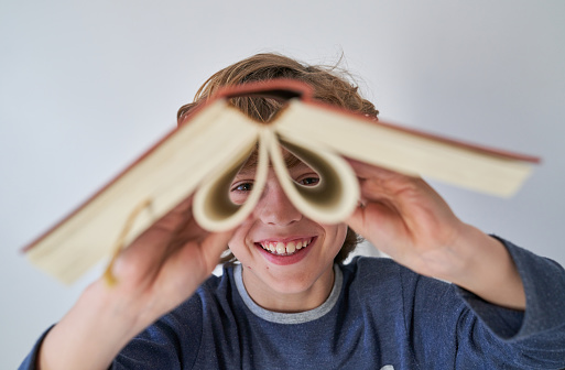 Happy preteen child in casual clothes smiling and making binocular from opened book while standing against light wall at home and looking at camera