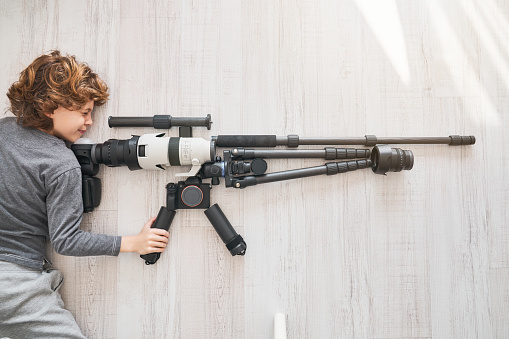 Top view of boy looking through scope of sniper rifle composed with equipment for photography while lying on wooden floor in light room during weekend day