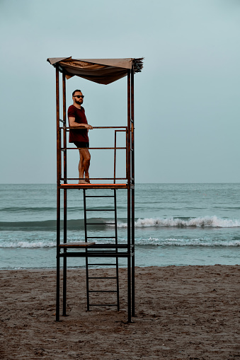 Male Lifeguard Standing On Tower And Keeping Watch Over Mediterranean Sea