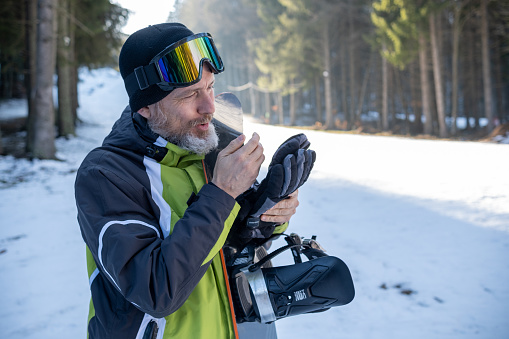 A cheerful elderly man with gray hair and a beard wearing a sports ski suit in green, black, gray, and white. On his head, he wears a black knitted hat and ski goggles. He adjusted his gloves. His snowboard rests against his chest. A sunlit ski slope can be seen in the background. He's trying to warm his hands with his breath!